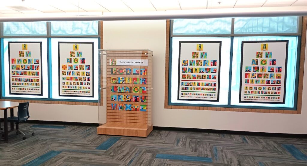 four art panels are hung on a wall in bright room, with a glass-enclosed case in the middle that displays "THE VISIBLE ALPHABET"