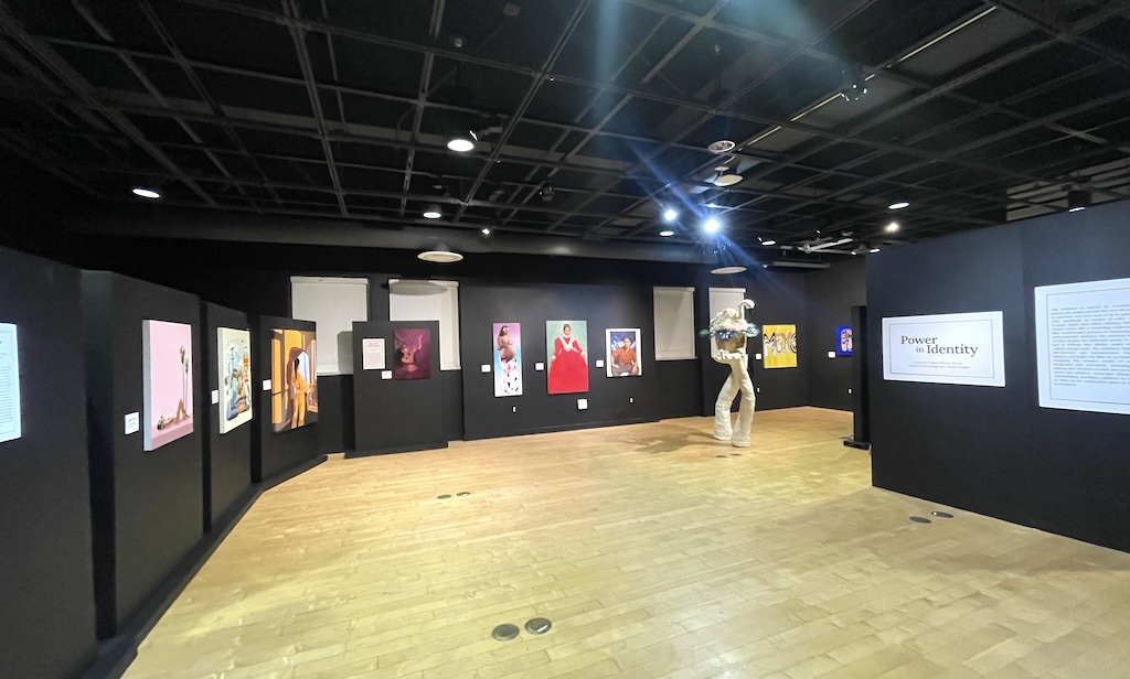 wide-angle photo of an exhibition space, with black walls surrounding a spacious open area, and a dozen pieces of art or panels on the walls.